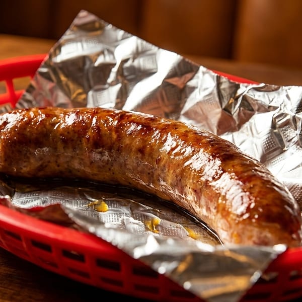 A glistening, cooked sausage rests in a red basket lined with crinkled aluminum foil on a wooden table.