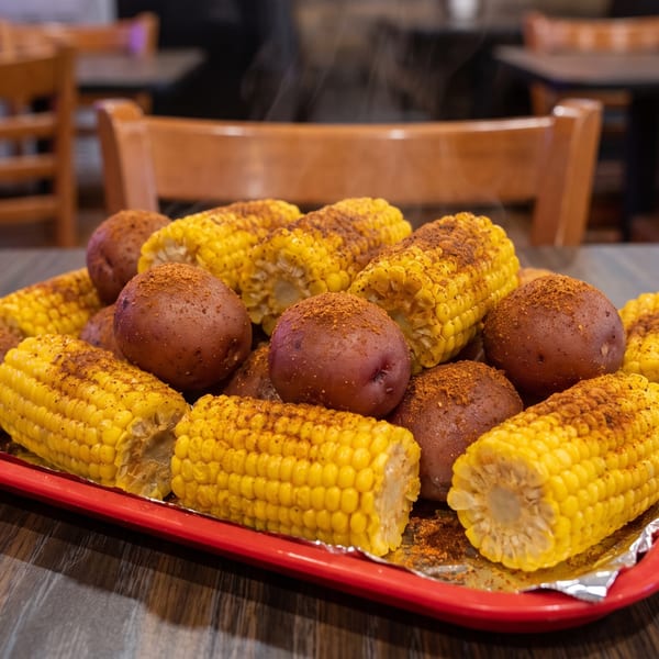 Steaming red potatoes and corn on the cob, seasoned with spices, on a red tray.