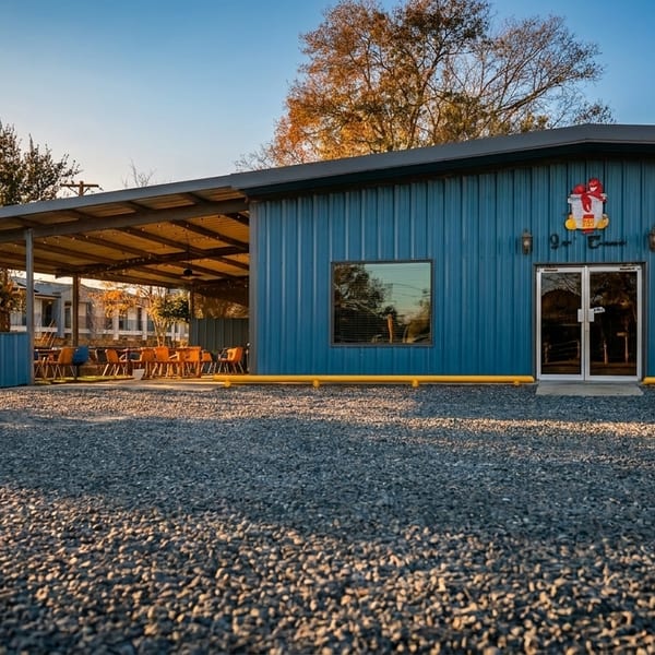 Blue corrugated metal building with a covered patio, gravel parking lot, and trees under a clear sky.