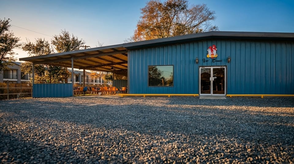 Blue corrugated metal building with a covered patio, gravel parking lot, and trees under a clear sky.